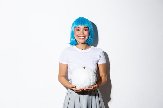 Beautiful asian girl in blue wig holding white pumpkin and smiling happy, celebrating halloween, standing over white background.