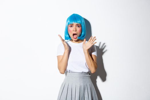 Image of horrified asian girl in blue wig screaming shocked, looking scared at something, standing over white background, dressed in halloween costume.