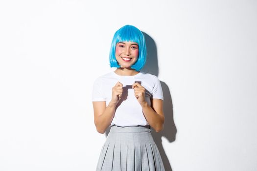 Smiling pretty asian girl showing credit card, wearing blue wig for halloween party, standing over white background.