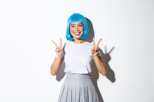 Cheerful asian girl in blue party wig, looking cute in halloween costume and showing peace gestures, standing kawaii over white background.