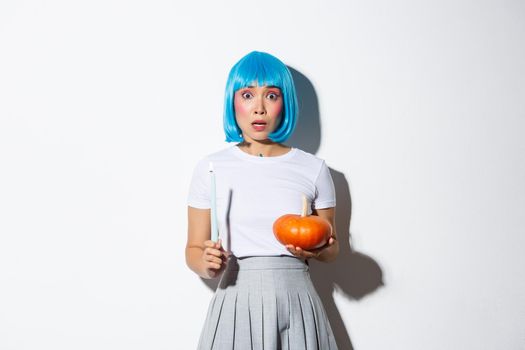 Concept of halloween. Image of scared asian girl in blue wig looking nervous and frightened, holding candle and pumpkin.