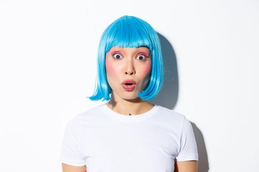 Image of cute asian girl in halloween costume and blue wig, looking surprised, standing over white background.