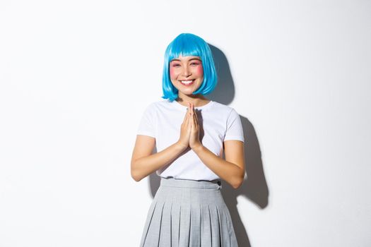 Portrait of lovely smiling asian girl in blue party wig, thanking for something, clasp hands together and looking grateful, standing over white background.