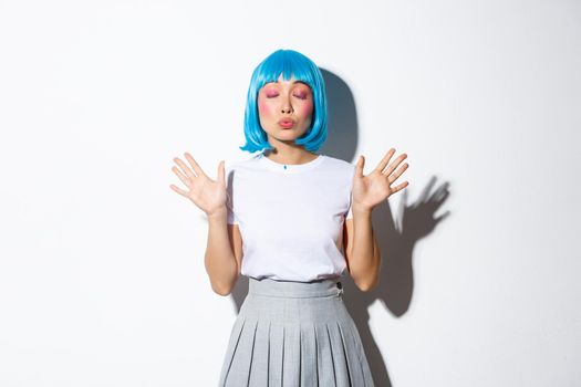 Portrait of cute asian girl in blue party wig, close eyes and pouting for kiss, standing over white background.