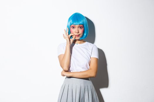Portrait of coquettish asian girl in blue wig seal her lips and promise not to tell secret, standing over white background.