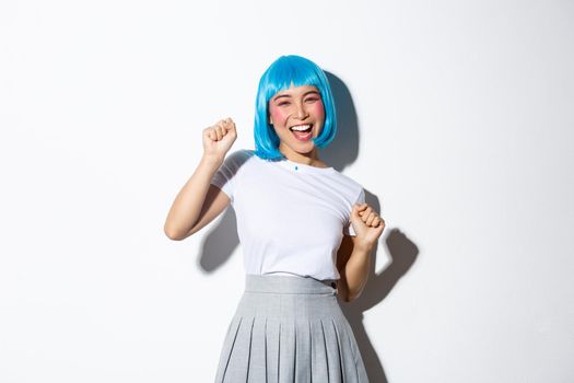 Portrait of carefree asian girl in blue wig having fun on party, celebrating halloween, dancing and laughing, standing over white background.