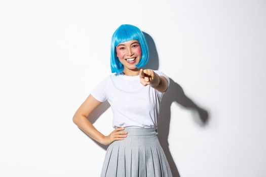 Portrait of happy beautiful asian girl smiling and pointing finger at camera, making choice, standing in blue wig over white background.