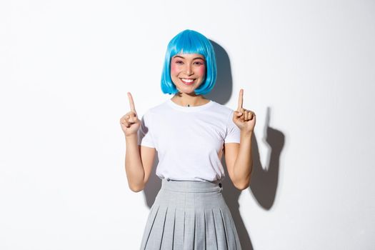 Cheerful asian woman in anime style outfit, wearing blue wig and smiling happy, pointing fingers up, showing advertisement, standing over white background.