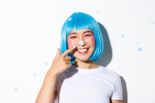 Image of cute and silly asian girl with confetti on her nose smiling and looking happy, wearing blue wig for halloween party.