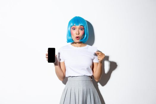 Portrait of surprised glamour girl in blue wig, gasping amazed, showing credit card and smartphone screen, standing over white background.