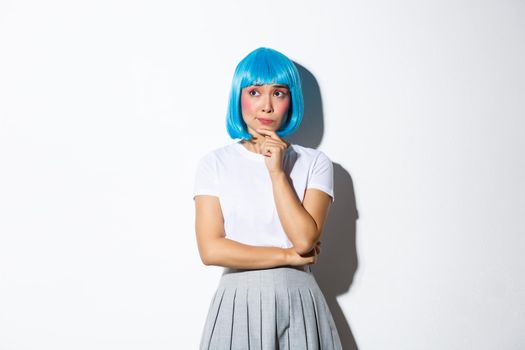 Image of troubled asian girl in blue short wig looking concerned and thoughtful, touching chin and gazing at upper left corner, thinking about something complicated, white background.