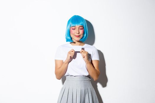 Portrait of beautiful asian girl in blue wig and halloween costume, daydreaming about shopping while holding credit card and smiling with closed eyes, standing over white background.