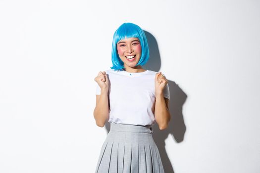 Portrait of cheerful asian girl celebrating victory, winning something and triumphing, standing in blue wig, celebrating halloween.