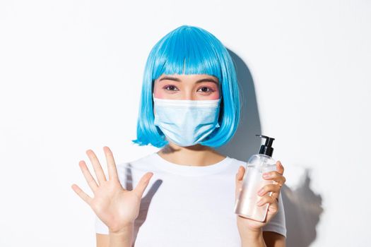 Concept of halloween celebration and coronavirus. Close-up of cute happy asian girl in medical mask and blue wig, showing clean hand and sanitizer, standing over white background.