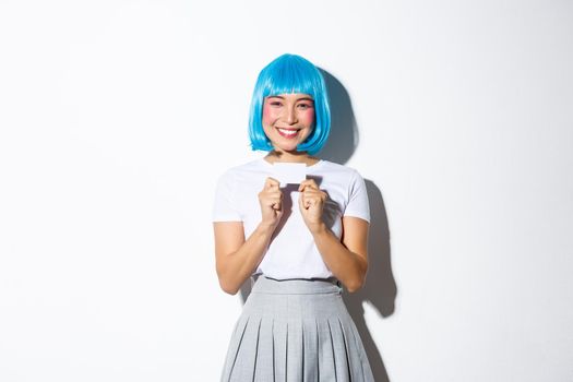 Portrait of cute asian girl in blue wig showing credit card, smiling happy, standing over white background.
