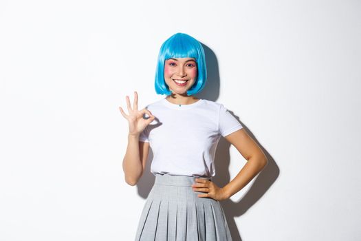 Portrait of smiling beautiful asian girl showing okay gesture in approval, recommend something good, standing in blue wig over white background.
