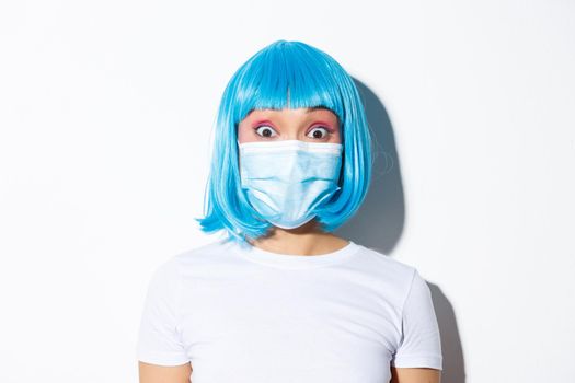 Concept of halloween celebration and coronavirus. Close-up of surprised asian girl in medical mask and blue wig, looking at camera amazed, standing over white background.