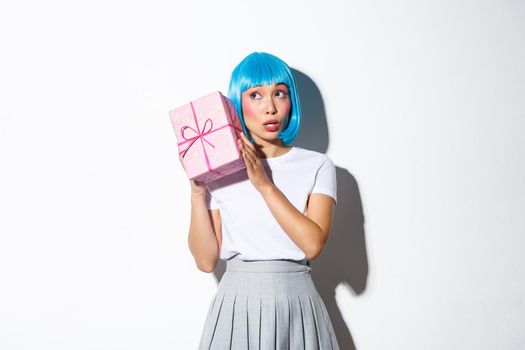 Cute asian girl in blue wig shaking box with gift, wonder whats inside, standing over white background.