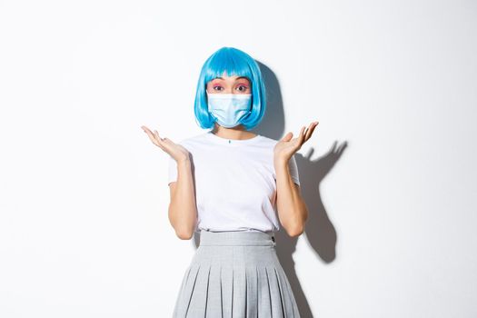 Concept of halloween during covid-19 pandemic. Portrait of surprised asian woman in blue wig and medical mask shrugging and looking amazed, standing over white background.