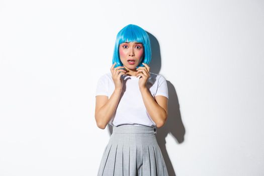 Portrait of scared asian woman in blue wig looking anxious and frightened, trembling from fear, wearing halloween costume, standing over white background.
