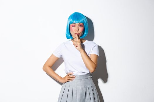 Sassy asian girl in blue wig share a secret, winking and shushing at camera with flirty expression, wearing blue short wig and halloween costume, white background.