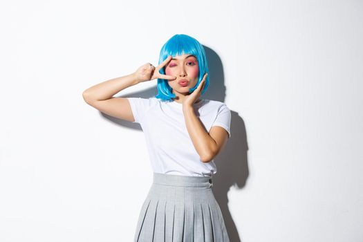 Image of flirty asian girl in blue anime wig showing peace gesture and winking at camera coquettish, standing over white background.