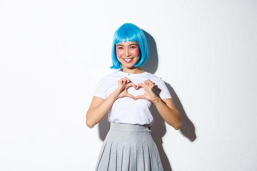 Portrait of lovely asian girl looking kawaii and showing heart gesture, smiling while standing over white background in halloween costume and blue wig.