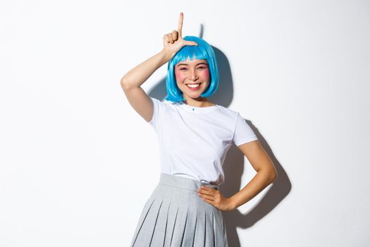 Portrait of beautiful silly asian girl celebrating halloween, showing loser gesture on forehead and smiling, mocking someone, standing over white background.