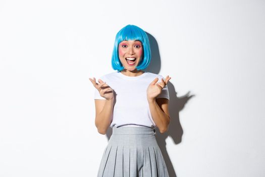 Portrait of happy and excited asian girl in blue short wig clap hands amused, smiling and looking at camera, standing over white background.