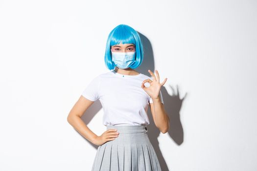 Concept of halloween celebration and covid-19 pandemic. Confident pretty asian female in blue wig and medical mask showing okay gesture.