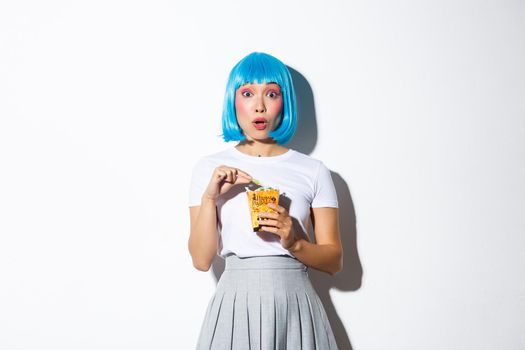 Surprised asian girl in blue wig and halloween costume holding bag with treats, standing over white background.