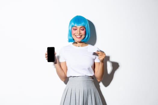 Beautiful smiling asian girl in blue wig looking pleased at credit card, showing smartphone screen, standing over white background.
