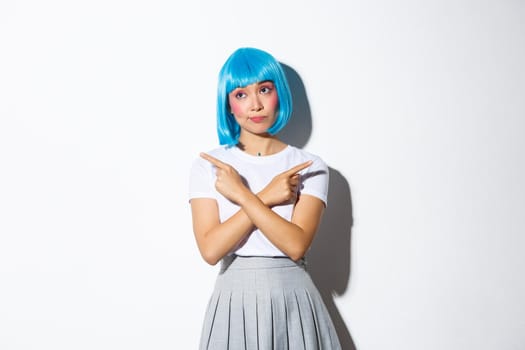 Indecisive beautiful asian girl in blue wig, pointing fingers sideways and looking puzzled while making choice, dressed for halloween, standing over white background.