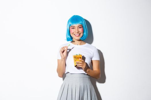 Portrait of cheerful asian girl in blue wig celebrating halloween, eating sweets from trick or treat bag, standing over white background.