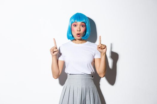 Surprised and fascinated asian girl in blue short wig, kawaii makeup, looking impressed and pointing fingers up, showing logo or promo banner, standing over white background.