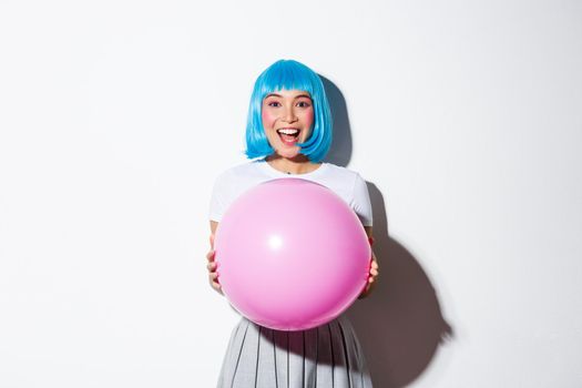 Image of cheerful asian girl in blue wig, celebrating holiday, wearing outfit for halloween party, holding large pink balloon and looking amazed.