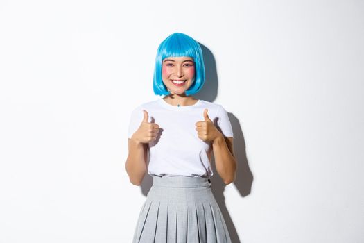 Portrait of satisfied smiling girl in blue wig and halloween costume, showing thumbs-up in approval, like something good, standing over white background.