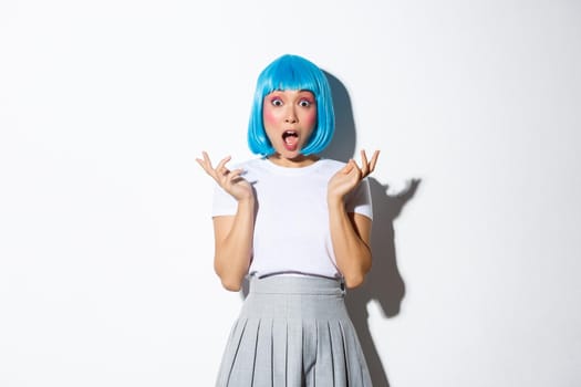 Portrait of surprised and excited asian party girl in blue wig looking amazed, spread hands and open mouth amazed as if hearing great news, standing over white background.
