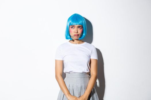 Portrait of complicated sad asian girl in blue wig, pouting and looking at upper left corner with troubled expression, standing over white background.
