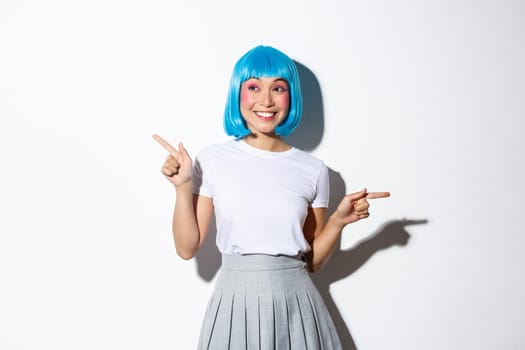 Portrait of amazed smiling woman in blue wig dressed for halloween celebration, pointing fingers sideways, showing two promos, standing over white background.