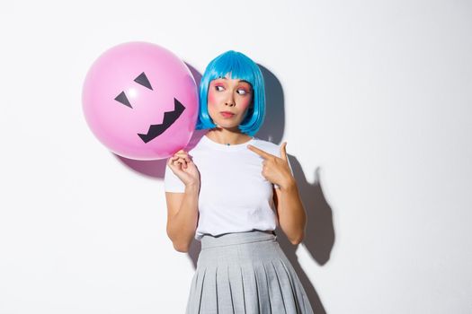 Beautiful asian girl celebrating halloween and wearing blue wig for party, pointing finger at balloon with scary face.