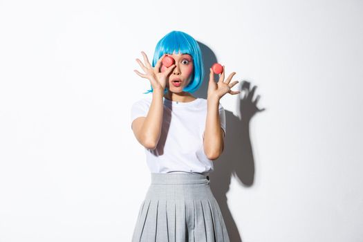 Portrait of surprised glamour girl in blue wig, gasping amazed, holding macaroons, standing over white background.