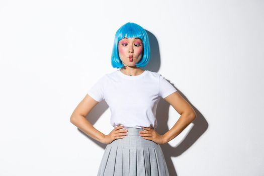 Image of funny cute asian woman in blue wig suking lips to make grimaces, joking around, dressed for halloween party or celebration event, standing over white background.