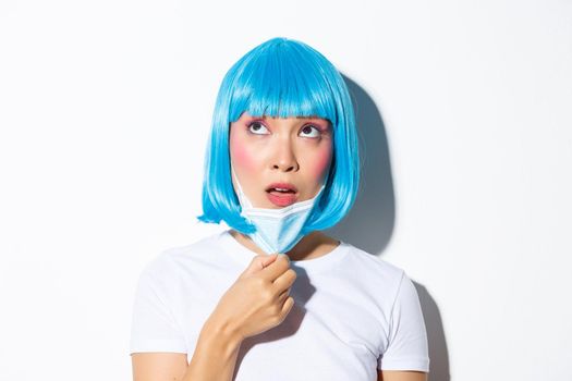 Concept of halloween celebration and coronavirus. Close-up of confused asian woman in blue wig, taking-off medical mask and looking up, standing over white background.