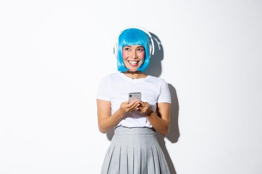 Image of coquettish asian girl listening music in wireless headphones, holding smartphone, looking up and smiling, standing over white background.