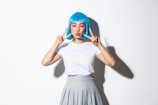 Image of kawaii asian woman in blue shirt wig, shows peace gesture and pout. Girl in halloween costume enjoys party, stands over white background.
