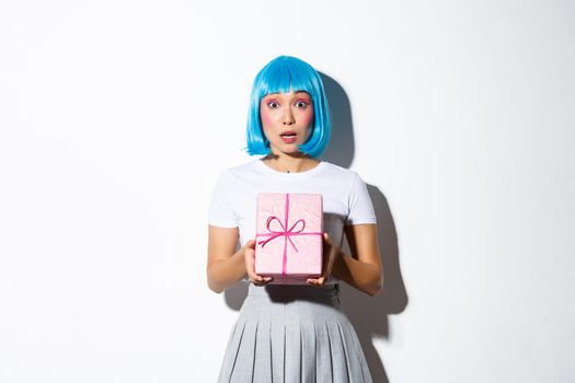 Surprised asian girl in blue party wig looking at camera confused, holding giftbox, standing over white background.