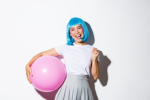Image of winning cheerful asian girl, looking happy and triumphing, celebrating holiday, wearing party outfit and blue wig, holding big pink balloon.