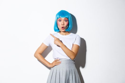 Curious attractive asian female in blue short wig and schoolgirl outfit pointing finger at upper left corner, looking intrigued, asking question about banner, standing over white background.