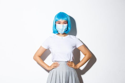 Image of cheerful asian girl ready for halloween party, protect herself from coronavirus as wearing medical mask, standing over white background in blue wig.